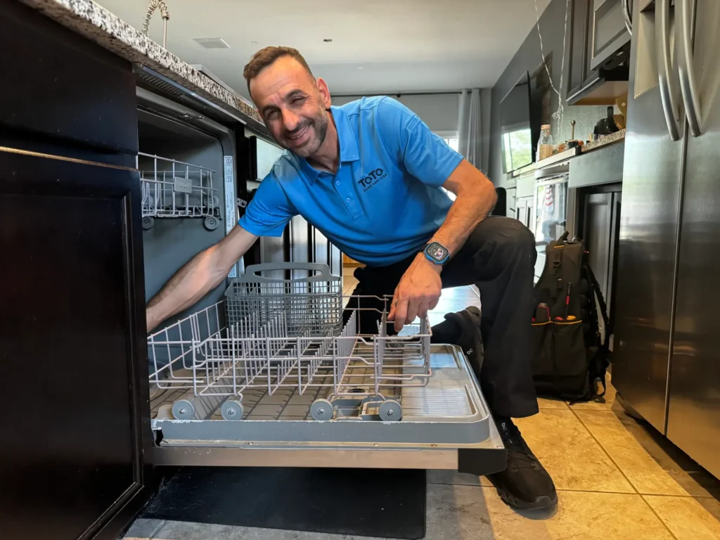 Man in a blue polo shirt kneels beside an open dishwasher, smiling while reaching inside the bottom rack in a modern kitchen.