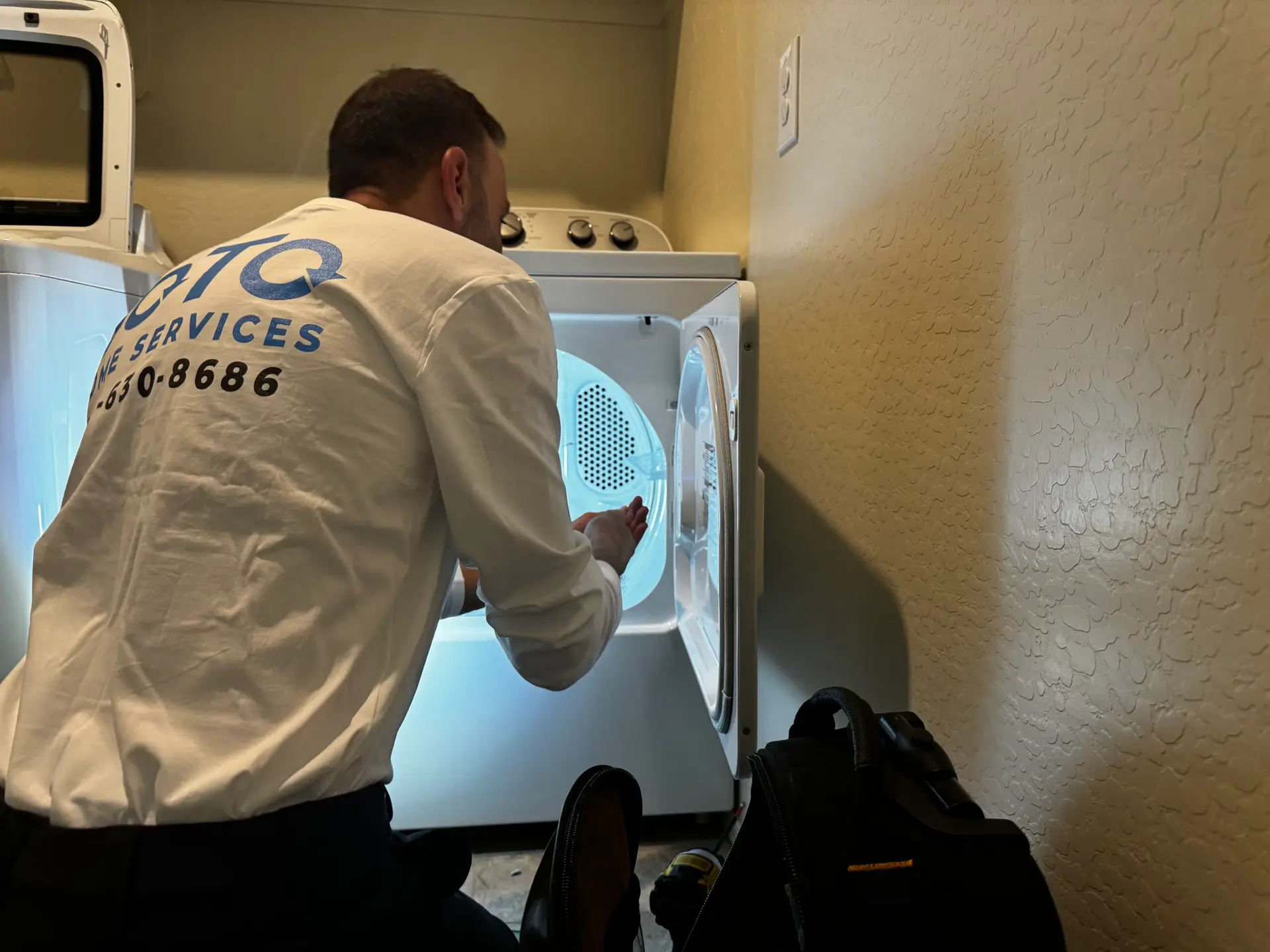 A technician kneels in front of an open clothes dryer, inspecting or repairing it, with a tool bag placed nearby on the floor.