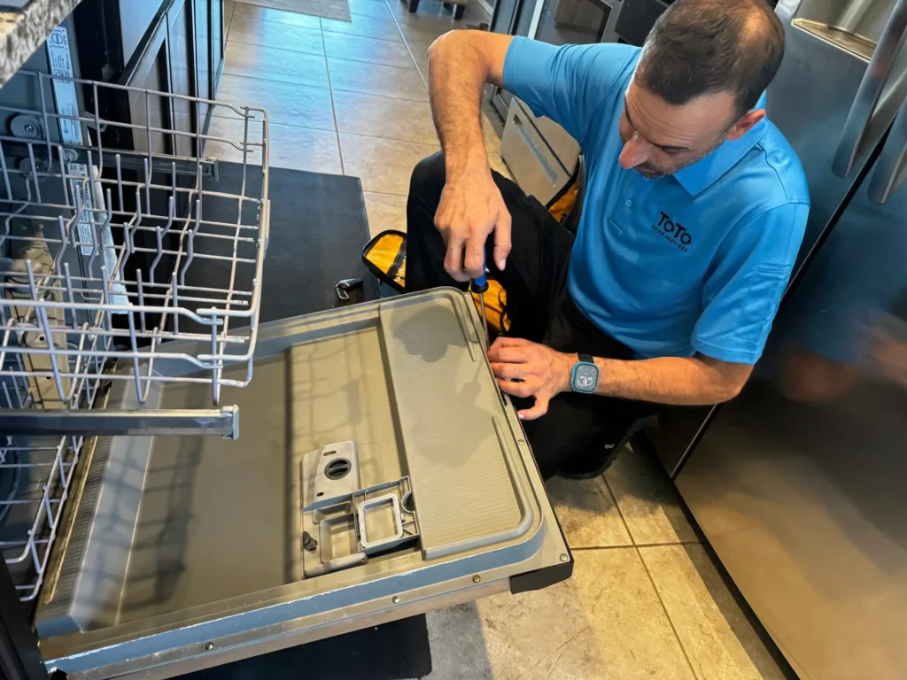 A technician in a blue shirt repairs an open dishwasher door using a screwdriver in a kitchen.
