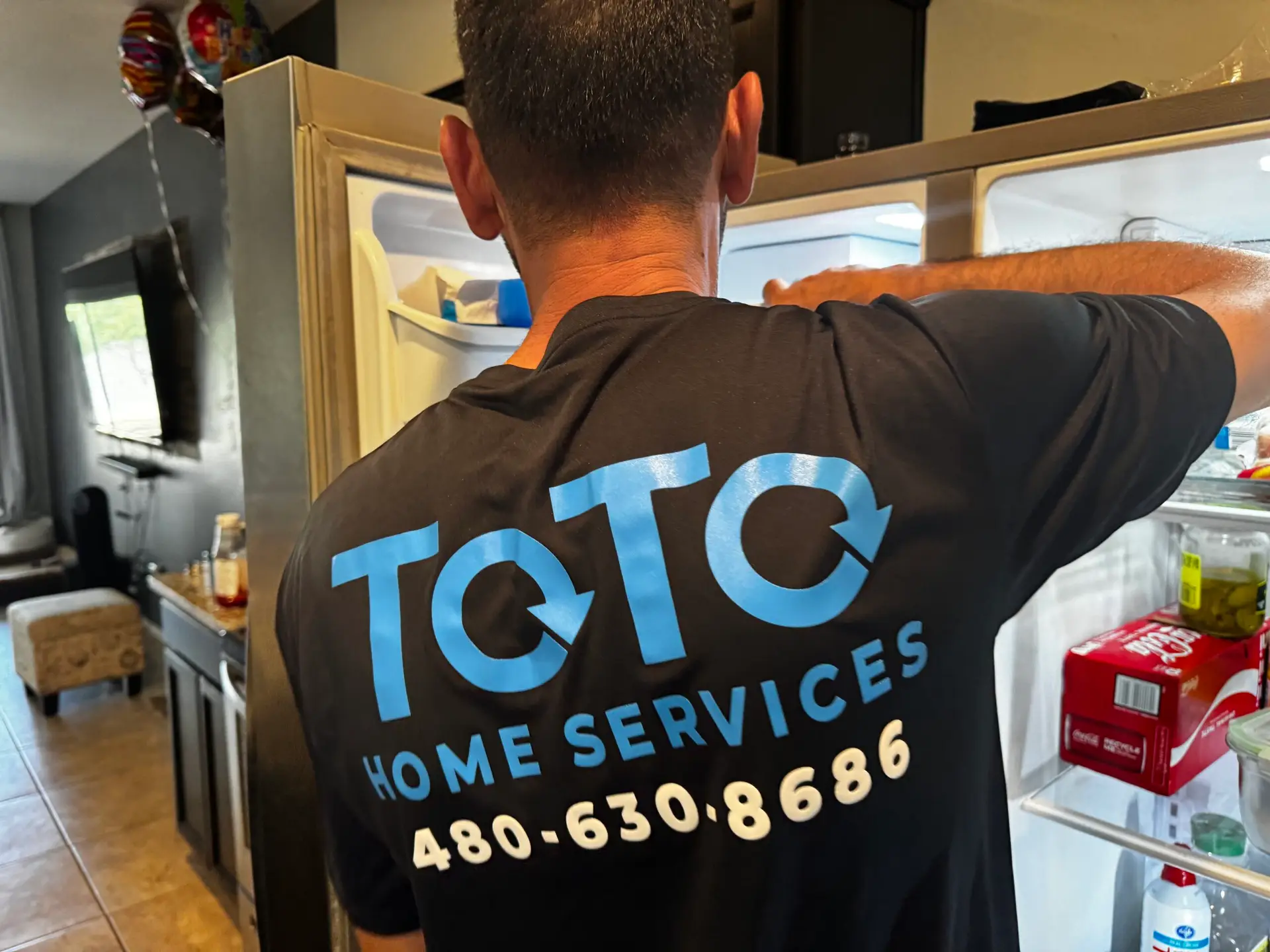 A person wearing a ToTo Home Services shirt with a phone number is standing in front of an open refrigerator in a kitchen.