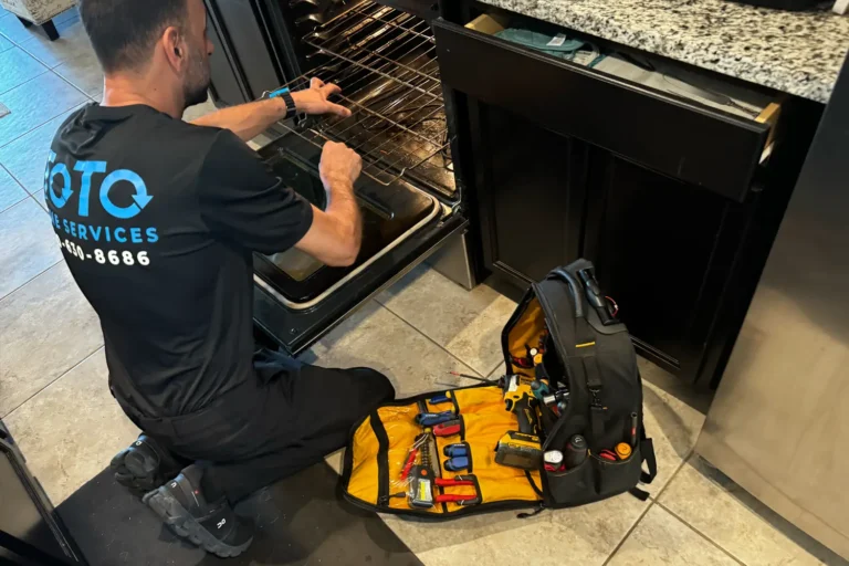 A technician kneels in front of an open oven, using tools for repair. A tool bag with various equipment is open on the kitchen floor beside him.