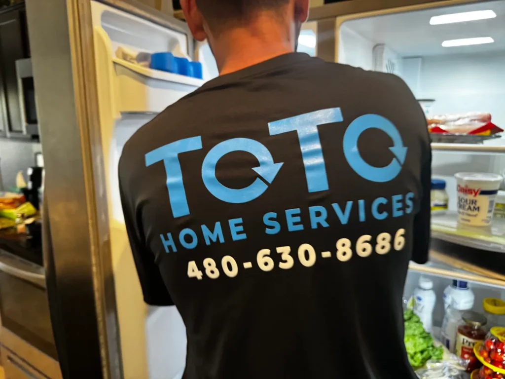 A person wearing a TOTO Home Services shirt with a phone number stands in front of an open refrigerator.