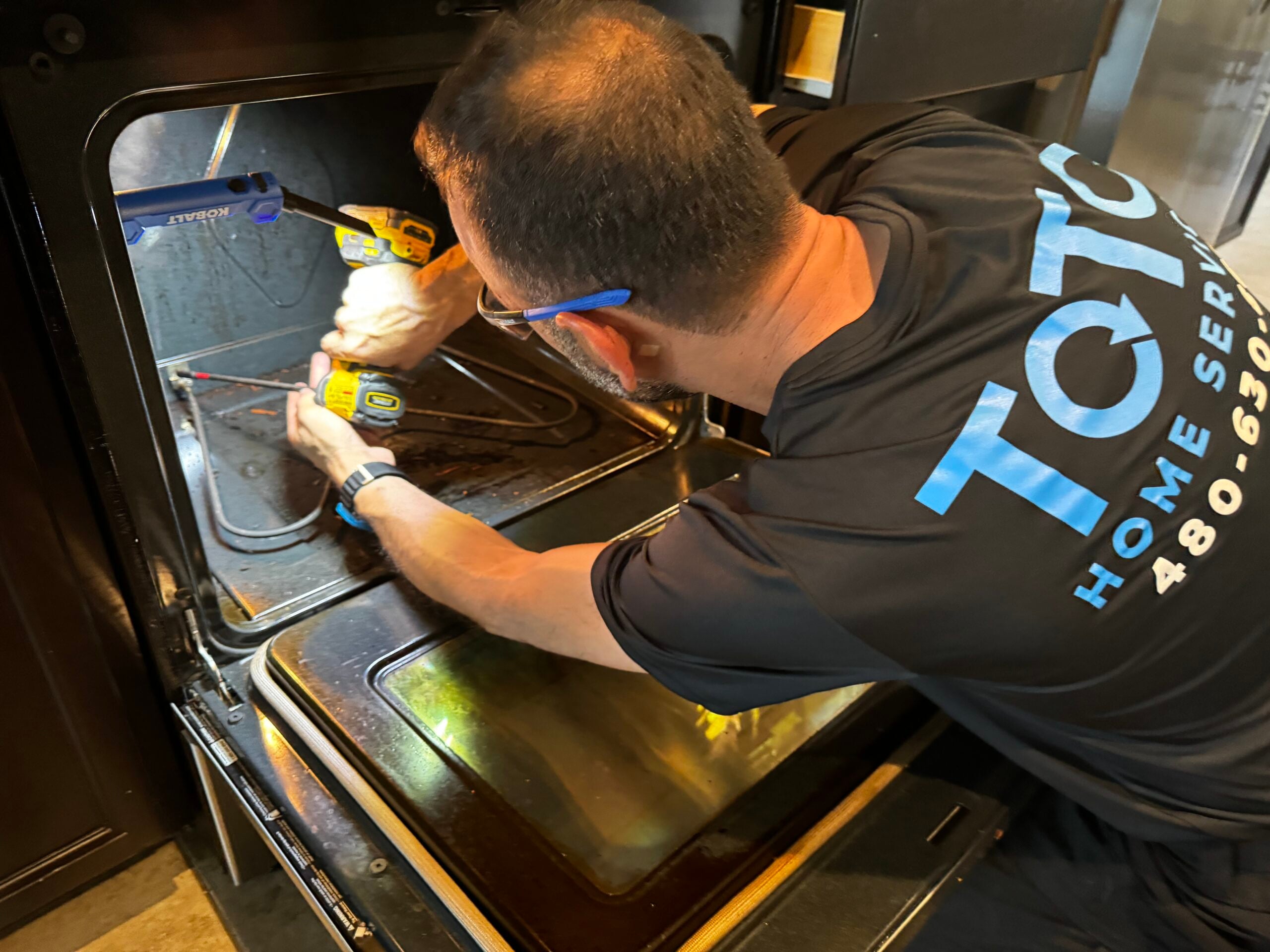 private-party-04 A technician in a TQTC Home Services shirt uses a power drill to perform maintenance inside an oven.