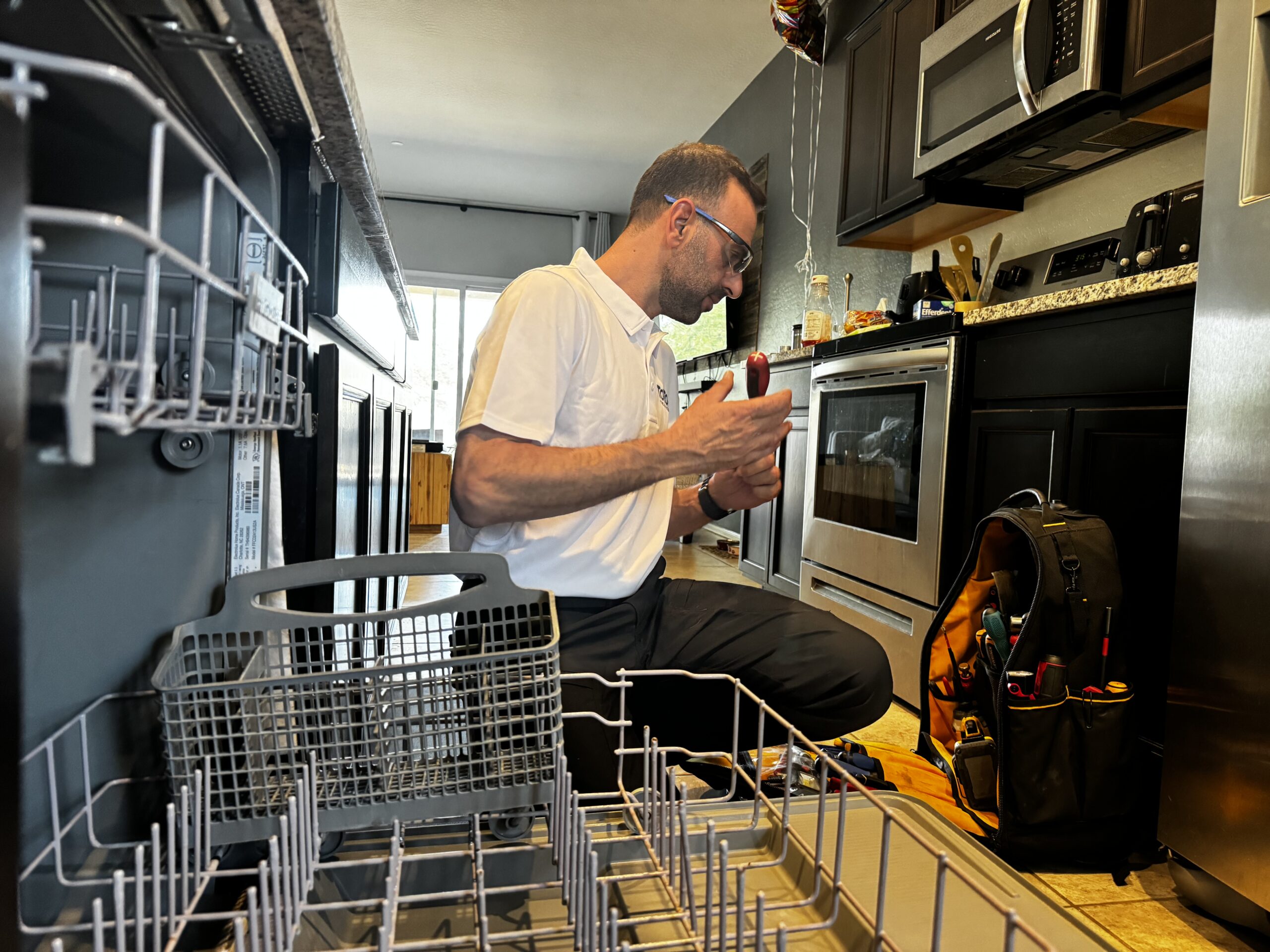 corporate-event-06 A technician kneels in a kitchen, examining a tool by an open dishwasher, with a tool bag nearby and kitchen appliances in the background.