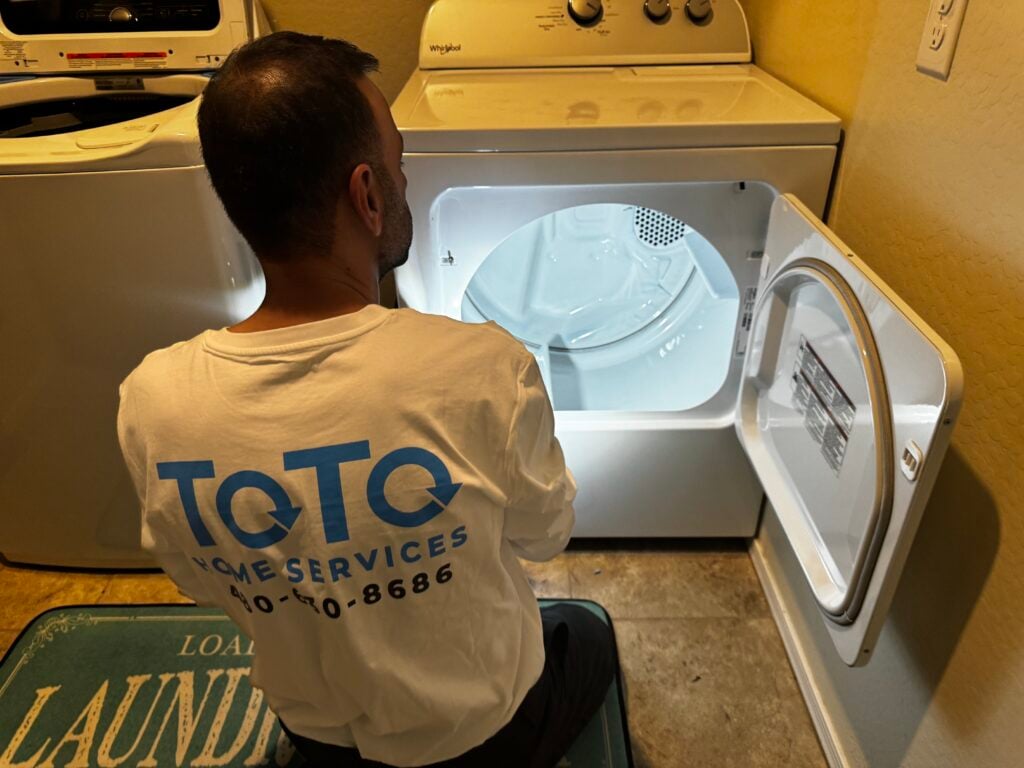 A man in a ToTo Home Services shirt kneels in front of an open dryer, inspecting its interior in a laundry room. A man in a ToTo Home Services shirt kneels in front of an open dryer, inspecting its interior in a laundry room.