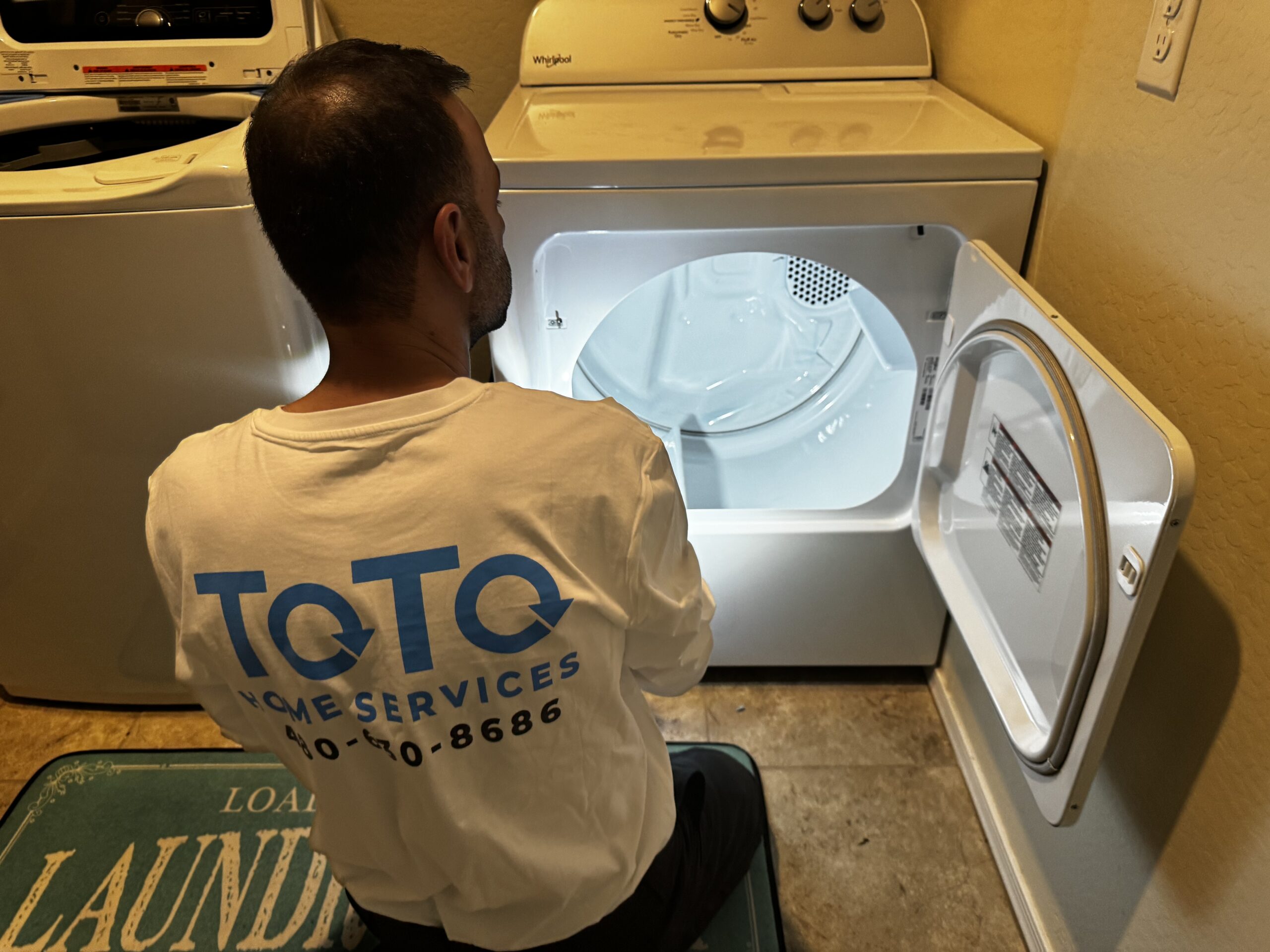 corporate-event-04 A man in a ToTo Home Services shirt kneels in front of an open dryer, inspecting its interior in a laundry room.