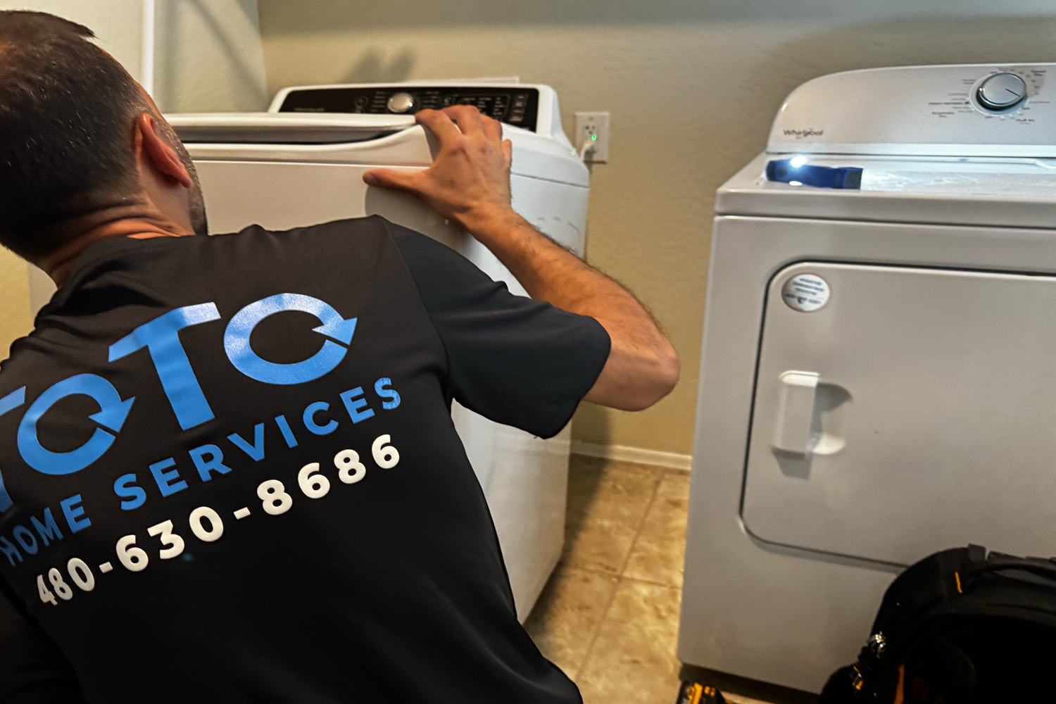 corporate-event-03 A technician from QTQ Home Services is repairing a top-load washing machine next to a white dryer in a laundry room.