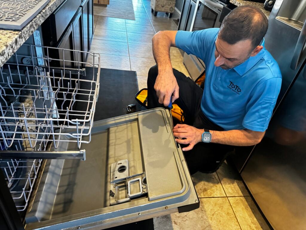 A technician in a blue shirt uses a screwdriver to repair the open door of a dishwasher in a kitchen. A technician in a blue shirt uses a screwdriver to repair the open door of a dishwasher in a kitchen.
