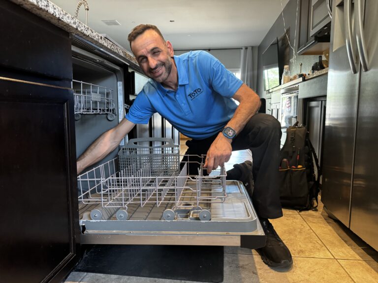 A man in a blue polo shirt kneels to inspect an open dishwasher in a modern kitchen.