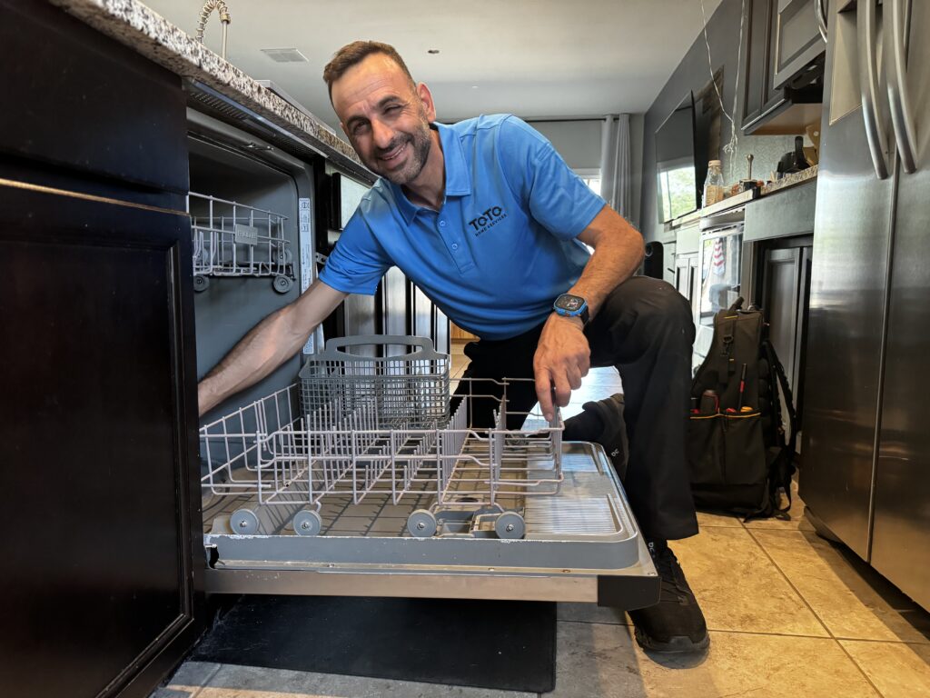 A man in a blue polo shirt kneels and inspects the lower rack of an open dishwasher in a modern kitchen.