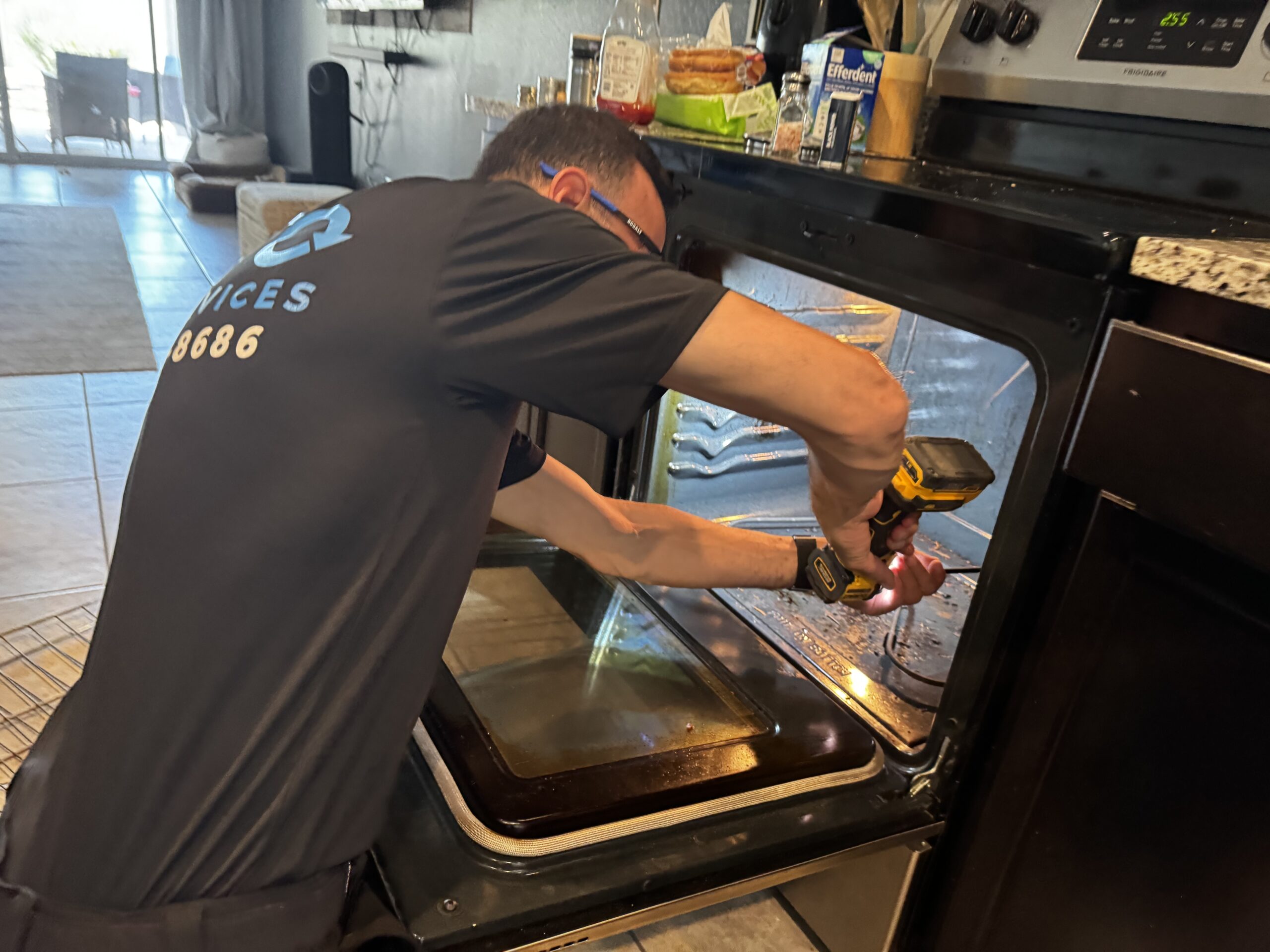 birthday-party-02 A man in work attire uses a power drill to repair or remove the door of an open oven in a kitchen.
