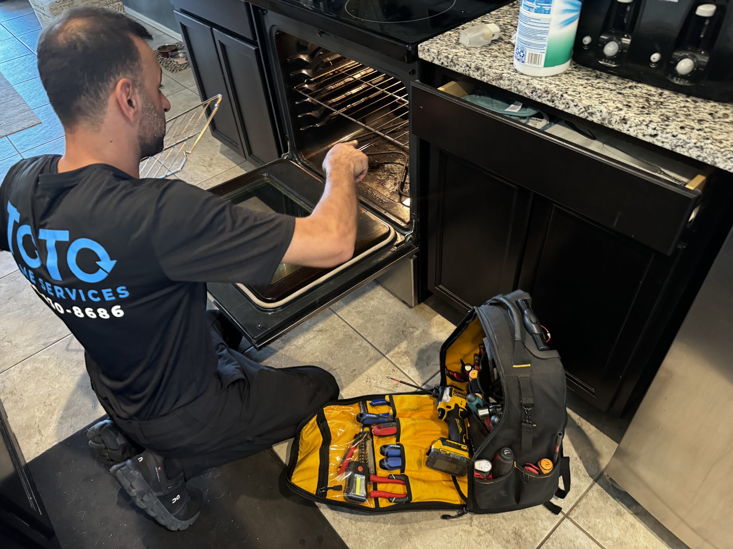 birthday-party-05 A technician kneels on the floor, inspecting an open oven with tools spread out from a yellow toolkit beside him in a kitchen.