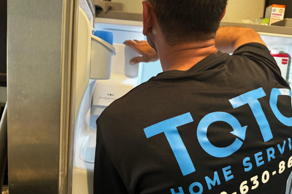 A technician wearing a ToTo Home Services shirt repairs the inside of a refrigerator. A technician wearing a ToTo Home Services shirt repairs the inside of a refrigerator.