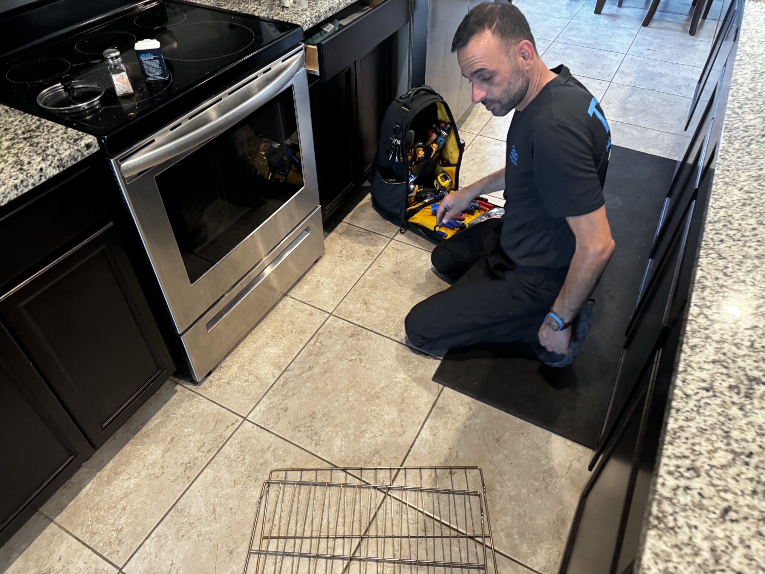 Oven Broiler Not Working — Is It the Element, the Igniter, or Something in the Control Board? A technician in a dark uniform kneels on a tile floor beside a stainless steel oven, holding a diagnostic device while an oven rack and tool bag are visible nearby.