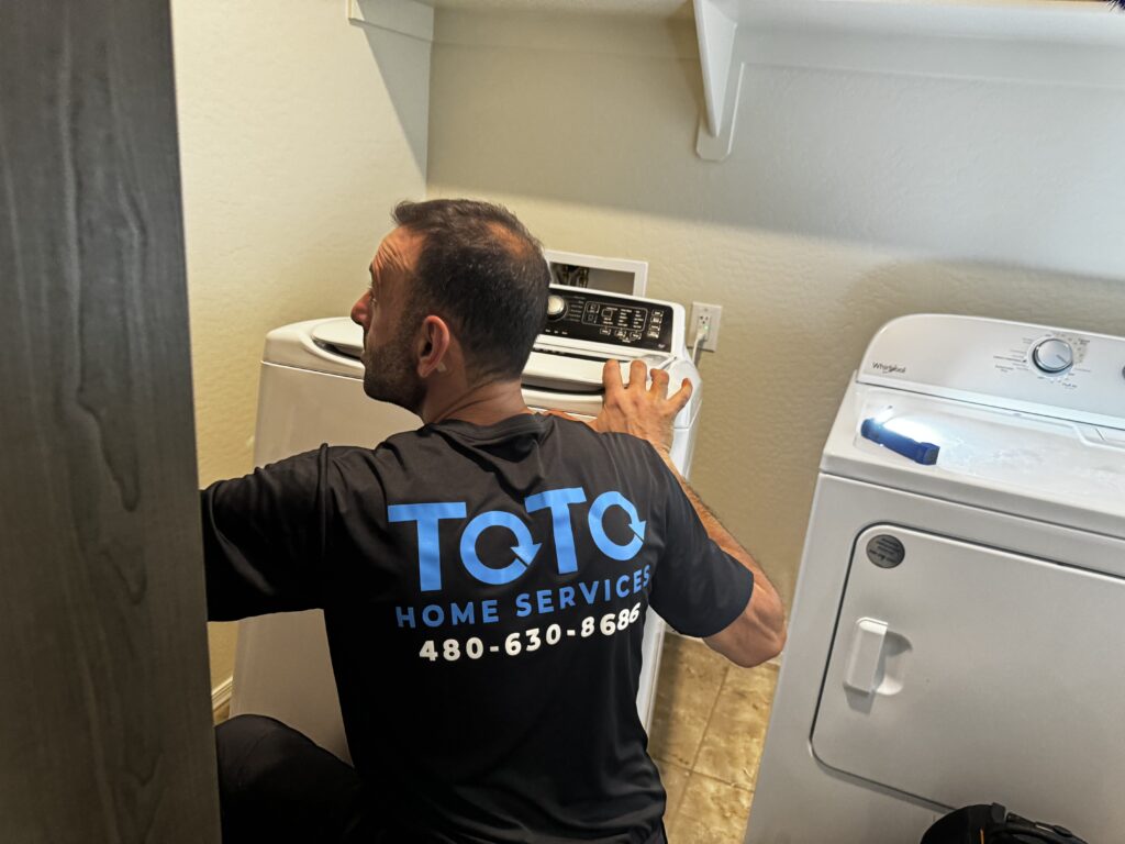 A technician wearing a TOTO Home Services shirt repairs a top-loading washing machine next to a dryer in a laundry room.